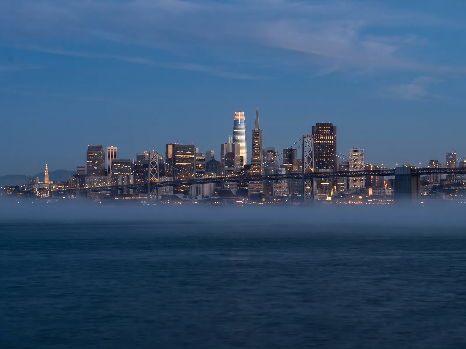 The downtown San Francisco skyline at dusk, seen across the bay with low fog on the water